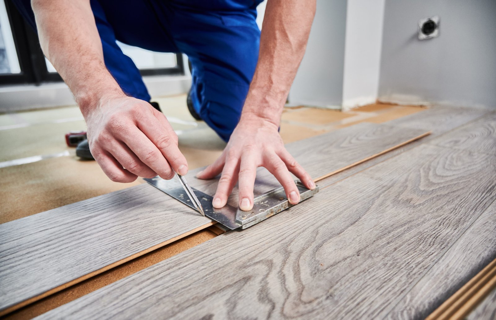 Close up of male worker using metal construction ruler and pen while drawing line on laminate flooring board. Man preparing laminate plank for floor installation in apartment under renovation.