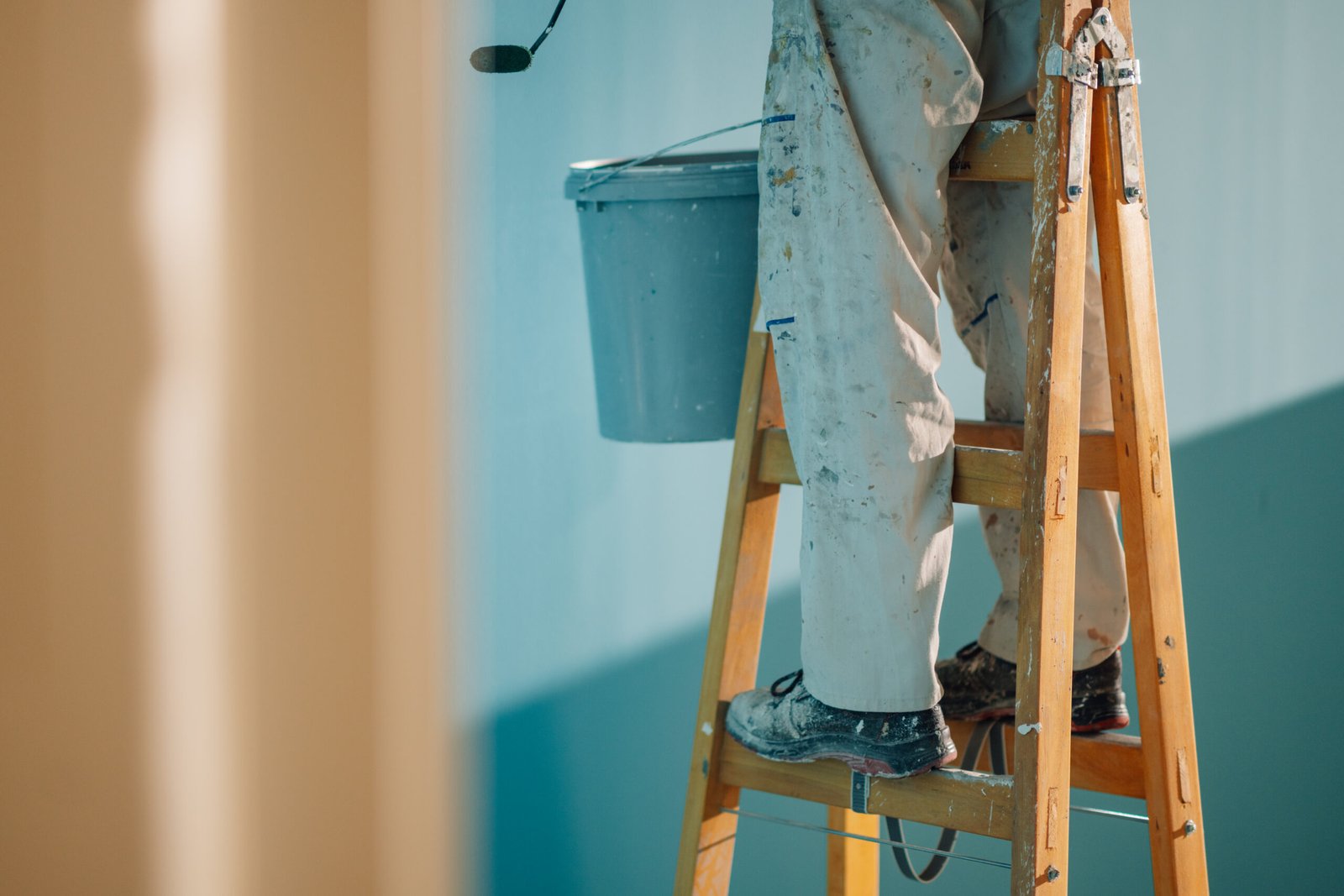 House painter standing on a wooden ladder, carefully painting a wall with a roller while a bucket of paint sits nearby, enhancing the interior